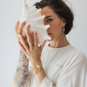 Frau, die eine große Muschel vor ihr Gesicht hält und dabei das oversized shirt trust in der Farbe ecru (ungefärbte Baumwolle) trägt.